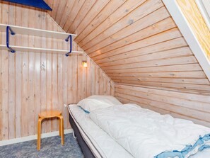 Una habitación con techo inclinado, paredes de madera, una cama con ropa blanca, un taburete de madera y una estantería con lámpara.
