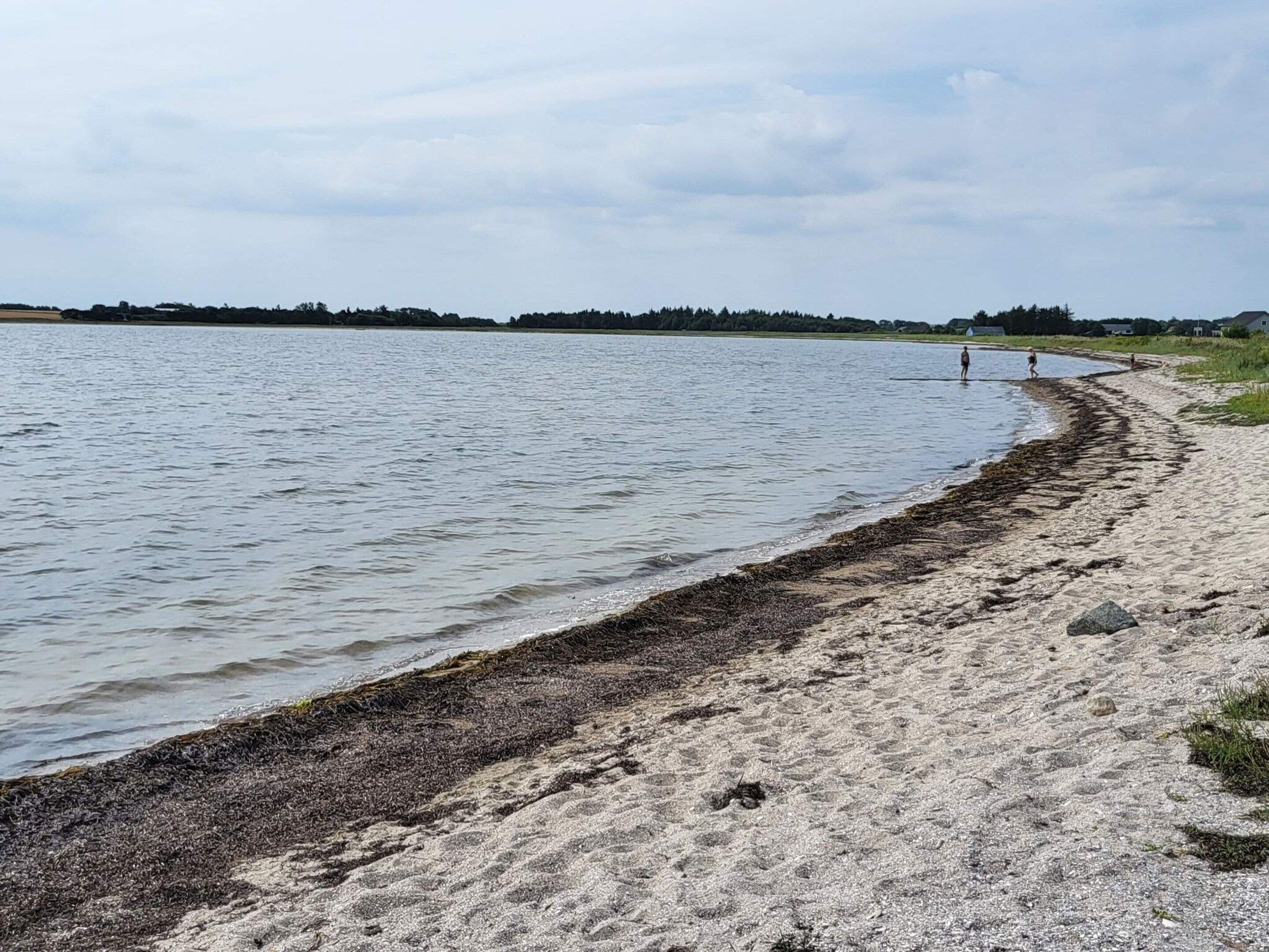 Une plage de sable fin bordée d’une étendue d’eau calme, quelques personnes qui se promènent et, au loin, une ligne d’arbres.