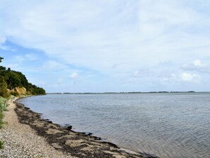 Paisaje costero con playa de guijarros, agua tranquila y una isla distante.
