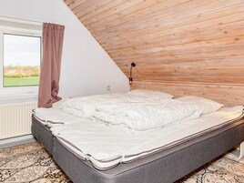 Un dormitorio con techo inclinado, una cama, un ventanal con vistas y una lámpara fijada a la pared.