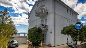 Un edificio di due piani con un cartello che indica che si tratta di un complesso residenziale. Sono presenti due balconi con ringhiere, un'auto parcheggiata davanti e piante in vasi intorno all'ingresso.
