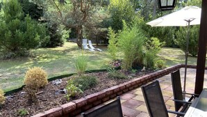 Un patio con borde de ladrillo, mobiliario exterior y vista a un jardín con plantas y árboles variados.