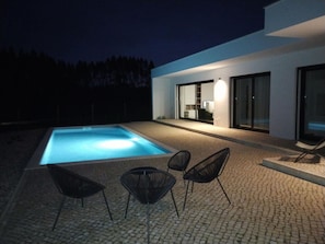 Uma área moderna de piscina ao ar livre, com piscina azul límpida, rodeada por um pátio pavimentado com quatro cadeiras pretas.