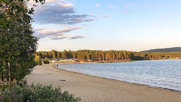 A beach with a sandy shore, a row of houses in the background, and a forested hill. - 8 Person Holiday Home in Osen (Åmot)
