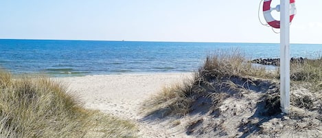 Une plage avec une bouée de sauvetage fixée à un poteau, une eau d’un bleu limpide et des dunes de sable.