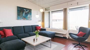 A modern living room with a dark grey sofa, a light-colored coffee table, and a blue armchair. There is a painting on the wall and a window with curtains.