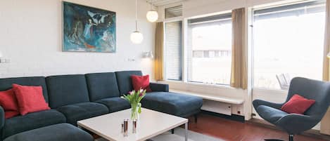 En moderne stue med en mørkegrå sofa, et lyst sofabord og en blå lænestol. Der er et billede på væggen og et vindue med gardiner.