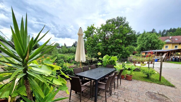 Une terrasse extérieure avec une table et des chaises, entourée d’une végétation luxuriante et protégée par un grand parasol.