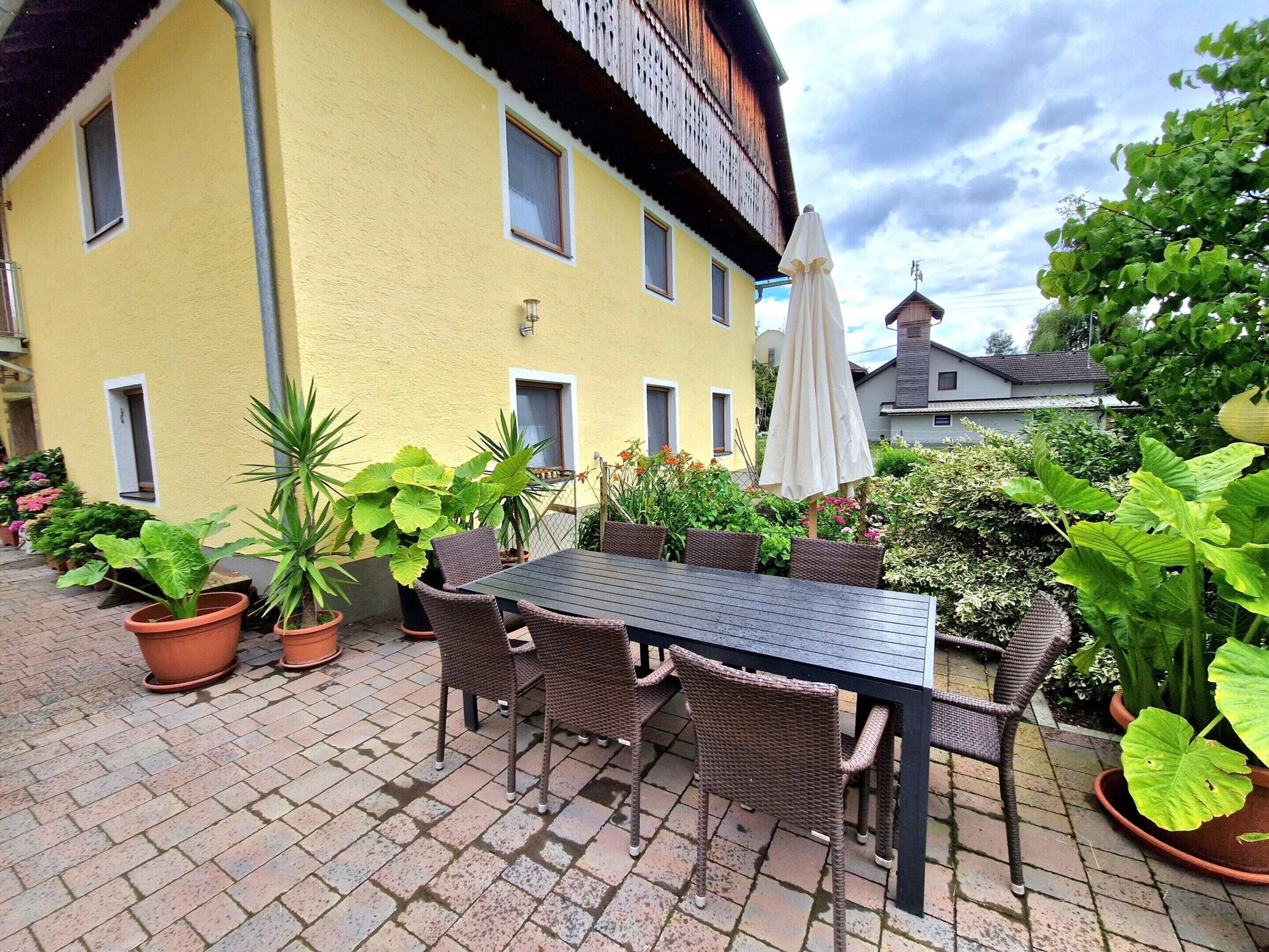An outdoor patio with a table and chairs, surrounded by potted plants and a yellow building in the background.
