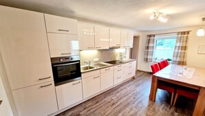 Une cuisine moderne avec des armoires blanches, un four encastré et un coin repas comprenant une table en bois et des chaises rouges.