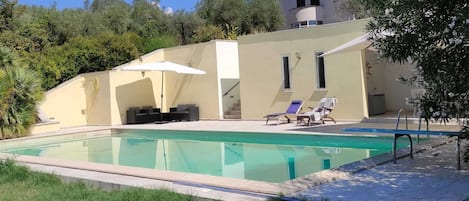 A swimming pool with lounge chairs and umbrellas, surrounded by greenery and a building with a balcony.