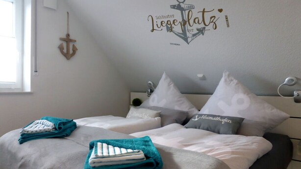 Un dormitorio con dos camas, cada una con toalla verde agua y blanca, un adorno de ancla en la pared y una ventana a la izquierda.