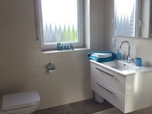 A bathroom with a toilet, a sink with a mirror, a window with a view of trees, and a towel rack with towels. - Inviting Apartment in Norden (Norden)
