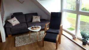 Una sala de estar con un sofá gris oscuro, una pequeña mesa blanca con una bandeja, una silla azul marino y un amplio ventanal con vistas a un jardín.