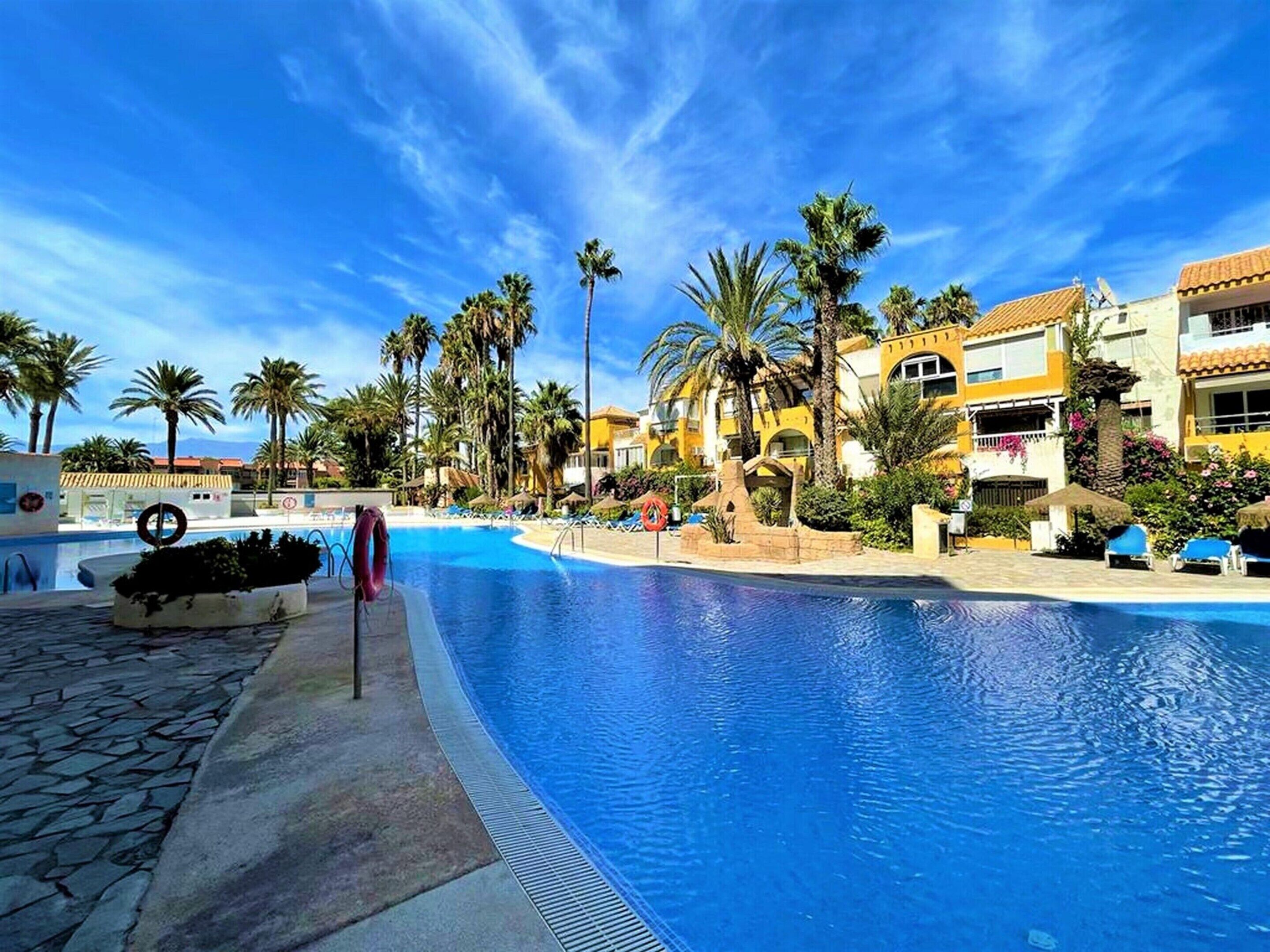 Una piscina con cielo azzurro, palme e un edificio dalle pareti giallo e bianco.