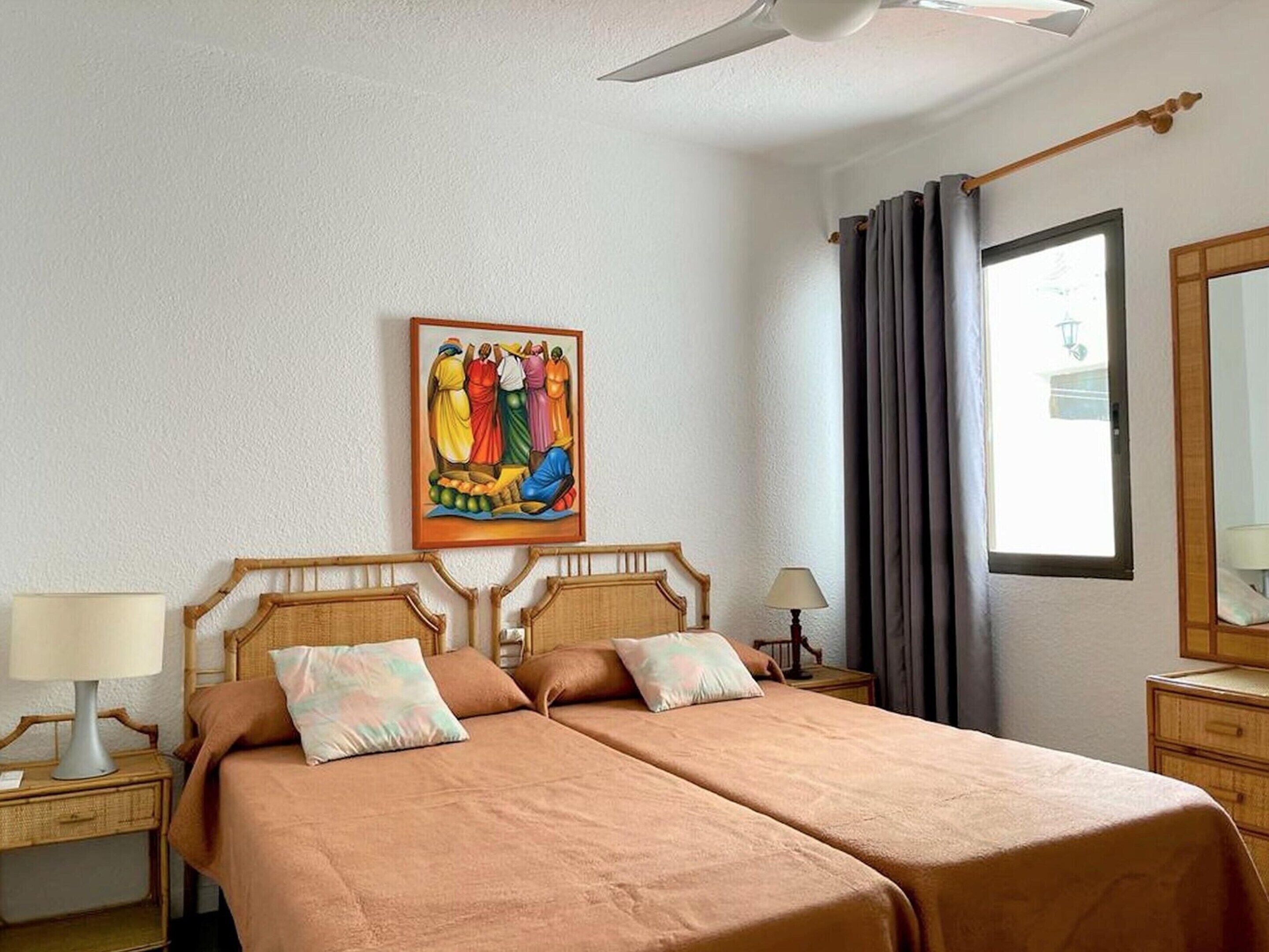 A double bed with a wooden headboard, a bedside table with a lamp, a painting on the wall, and a window with curtains.
