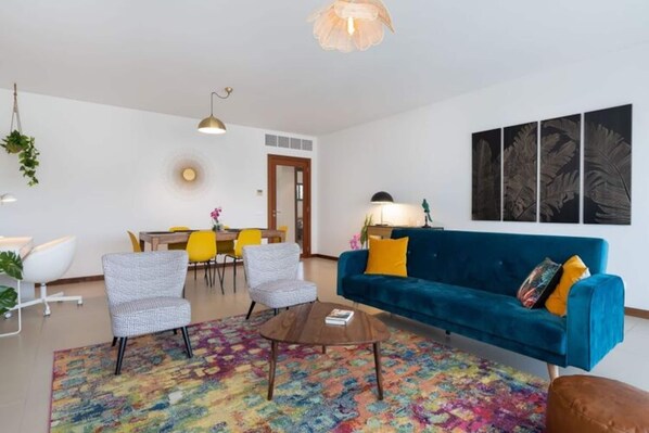 A modern living room with a blue sofa, two armchairs, a coffee table, and a colorful rug. There is a dining area with a yellow table and chairs, and a wall with framed artwork. - Apartment Marina Sky (Lagos)