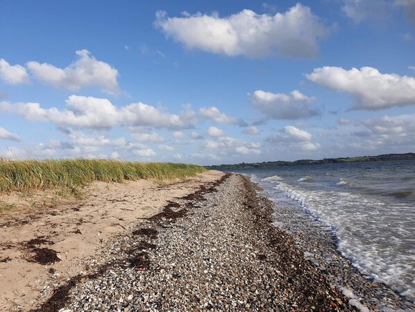 En strand med smÄsten, en grÀsklÀdd sanddyn och en klar himmel.