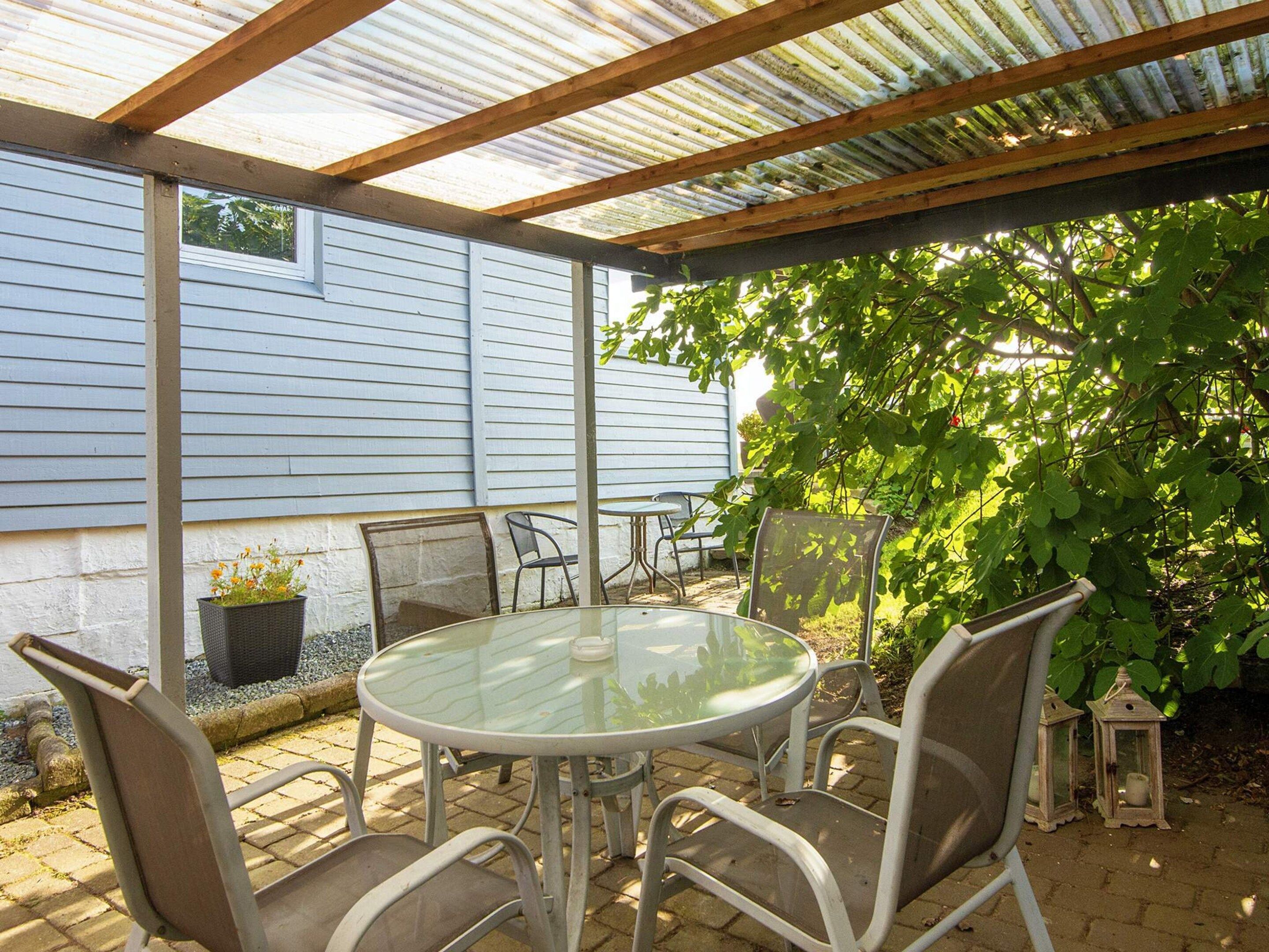 An outdoor patio with a round glass table and metal chairs, a covered area with a metal roof, and a house in the background.