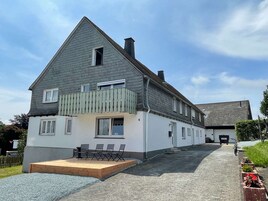 Uma casa de dois andares com varanda, um alpendre com cadeiras e uma entrada de automóveis que leva a uma garagem.