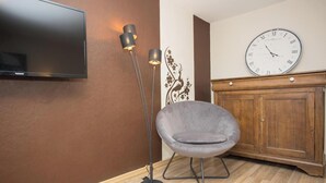 Ein Zimmer mit Flachbildfernseher, einem grauen Sessel, einer hohen Stehlampe, einem Holzschrank und einer Wanduhr.