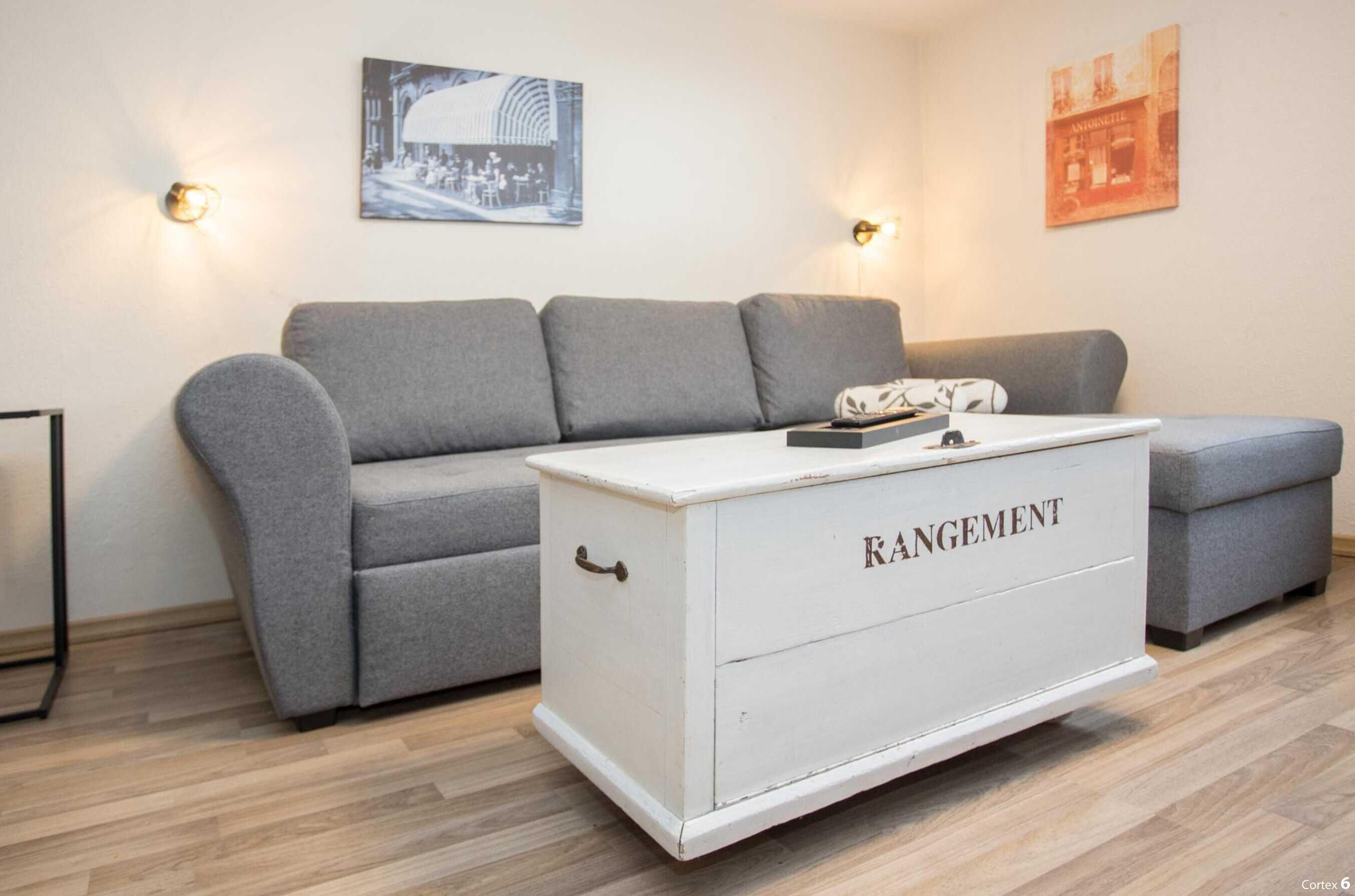 A living room with a grey sofa, a white chest of drawers labeled "RANGEMENT," and two wall-mounted light fixtures.