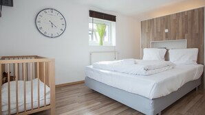 Ein Schlafzimmer mit einem Holckrippenbett, einem Bett mit weißer Bettwäsche, einer Wanduhr und einem Fenster mit Jalousien.
