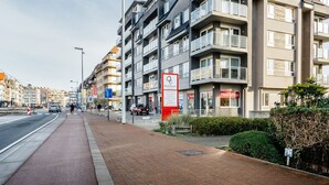 Een meerhoog woongebouw met balkons, een voetpad en een straat met auto's en een zebrapad.