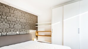 Een slaapkamer met een groot bed, een behangetje met een patroon als hoofdbord, een kledingkast met open planken, en een nachtkastje met een lamp.