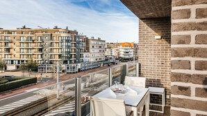 Een balkon met een tafel en stoelen, met uitzicht op een straat met gebouwen en een tram.