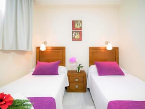 A double room with two beds, each with a wooden headboard and white bedding. There are two bedside tables with lamps, a small nightstand with a vase of flowers, and a framed picture on the wall.