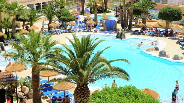 A resort pool area with palm trees, lounge chairs, and a large building in the background. - Belvilla by OYO Apartamento 2 Dormitorios (Roquetas de Mar)