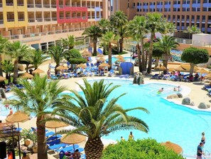 Zona de piscina en un resort, con palmeras, sillones de descanso y un edificio grande al fondo.