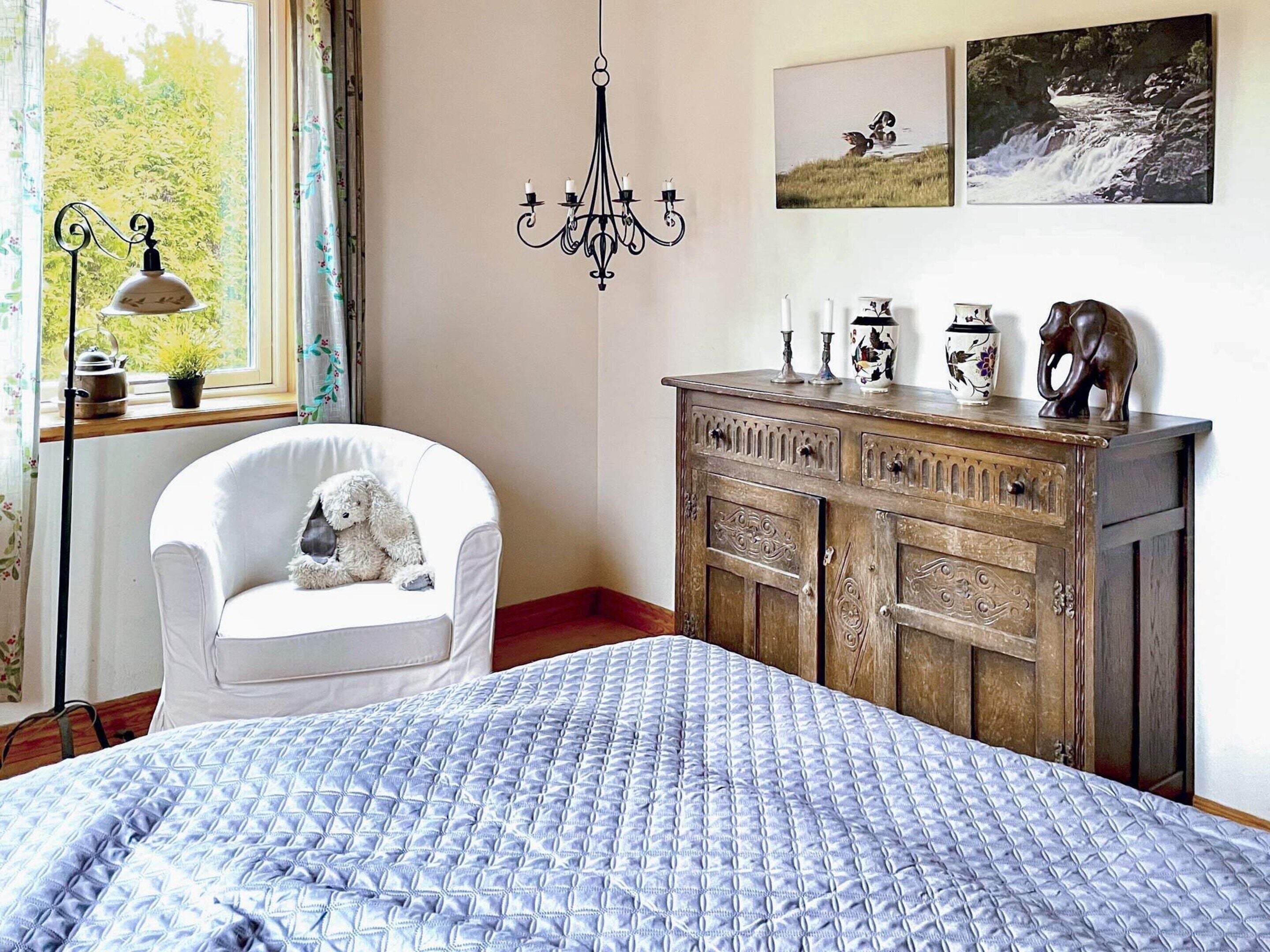 A bedroom with a bed, a white armchair, a wooden dresser, a chandelier, and two framed pictures on the wall.