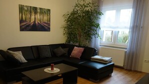 A living room with a black sofa, a coffee table, a large window with curtains, and a painting of a forest on the wall. - Elisabeth on the Loreley (Sankt Goar)