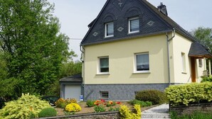 A two-story house with a stone foundation, a grey roof, and a light-colored exterior. There is a driveway with a stone wall and a small garden with various plants and flowers. - Elisabeth on the Loreley (Sankt Goar)
