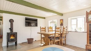 A cozy room with a wood-burning stove, a dining table with chairs, a TV mounted on the wall, and a window with a view.