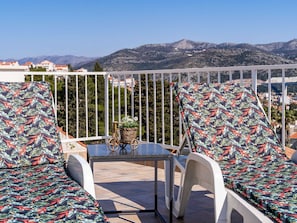 A balcony with two white chairs and a table, a wicker basket with a plant, and a view of mountains and buildings in the background. - Apartment Dorica - Two Bedroom Apartment With Terrace (Dubrovnik)