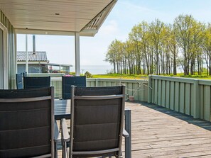 A covered outdoor deck with two chairs and a table, overlooking a grassy area with trees and a body of water. - 5 Person Holiday Home in Juelsminde (Horsens)