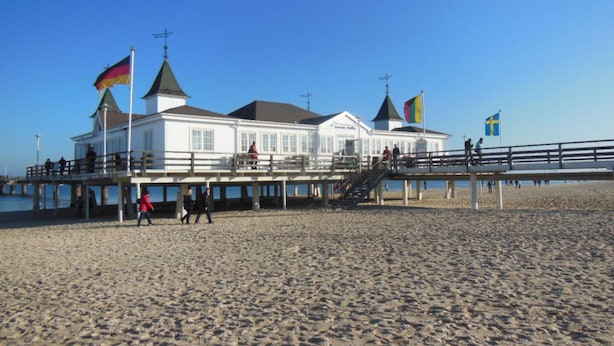 Un bâtiment situé au bord de la plage, doté d’une jetée, de plusieurs drapeaux et d’un escalier menant à l’étage supérieur.