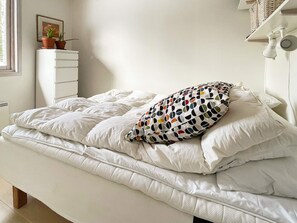 A bed with white bedding and a multicolored pillow, a white bedside table with a plant, and a wall-mounted shelf with baskets. - 6 Person Holiday Home in Blido (Blidö)