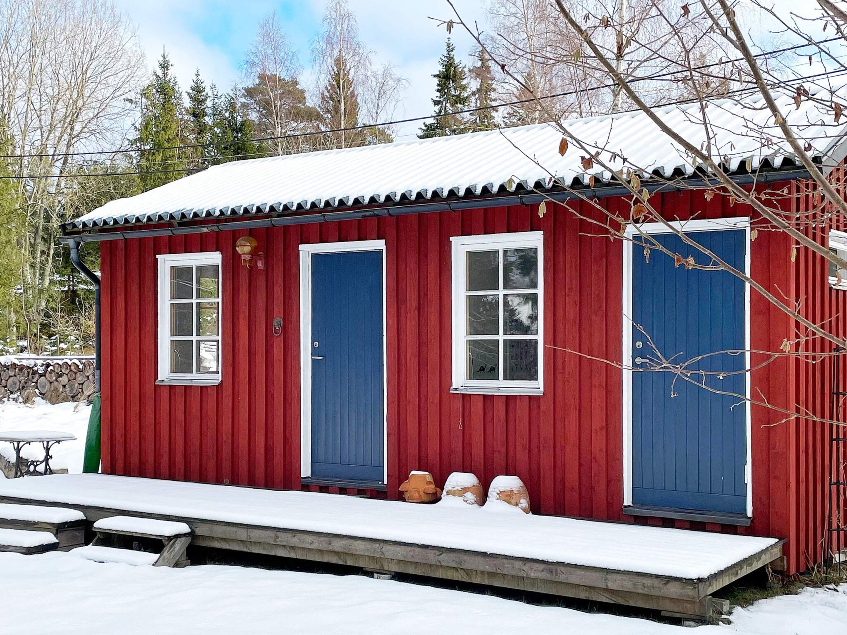 A red wooden cabin with two blue doors and windows, a small porch, and a snow-covered ground.