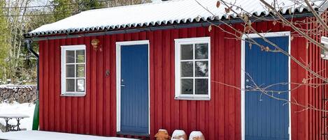 A red wooden cabin with two blue doors and windows, a small porch, and a snow-covered ground.