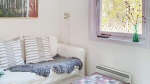 Une pièce avec un canapé blanc, une petite table sur laquelle est posée une courtepointe, un tableau au mur et une fenêtre donnant sur des arbres.
