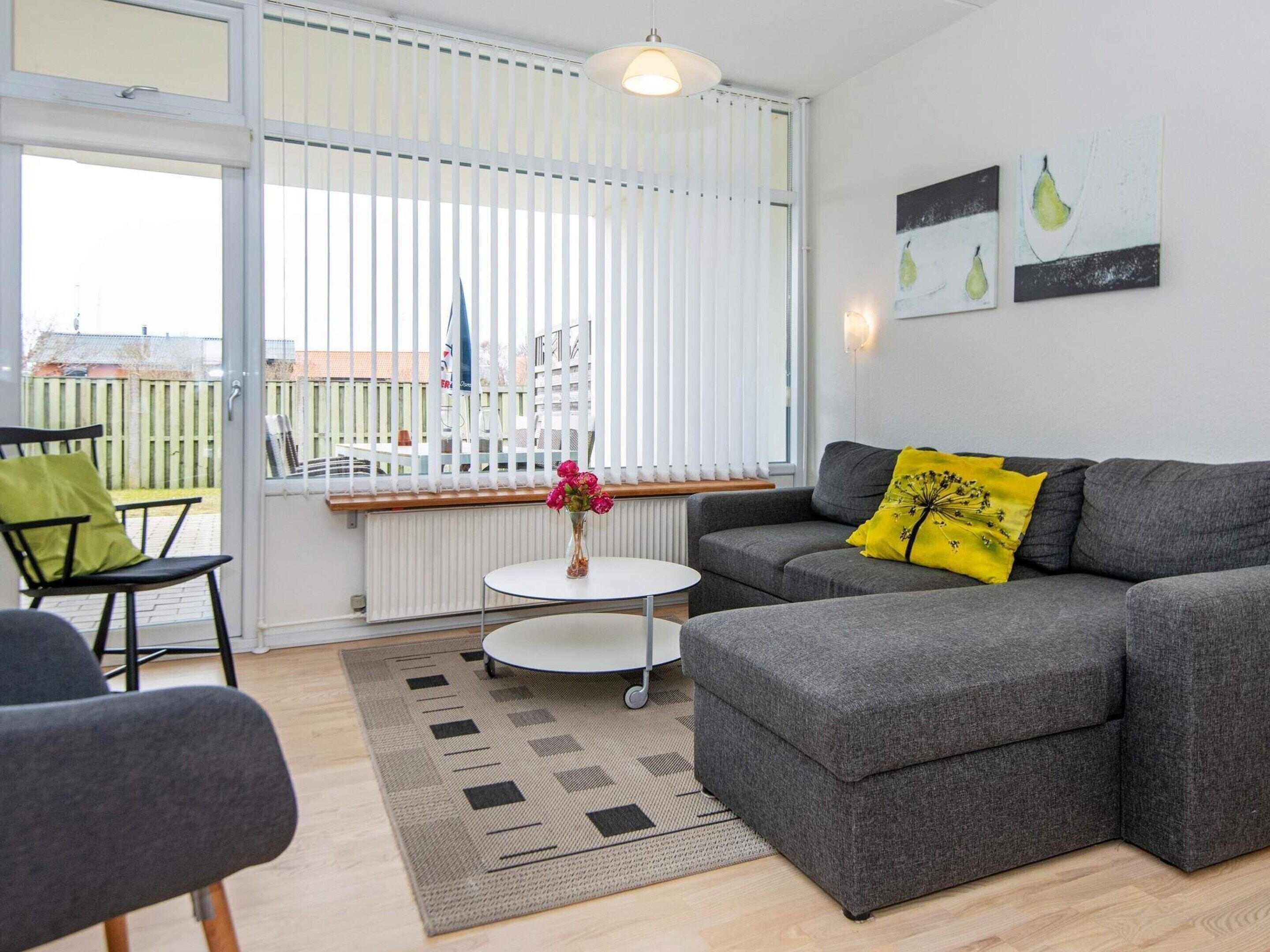A modern living room with a grey sofa, a small round table, and a chair. There is a window with blinds, a radiator, and a yellow pillow on the sofa.