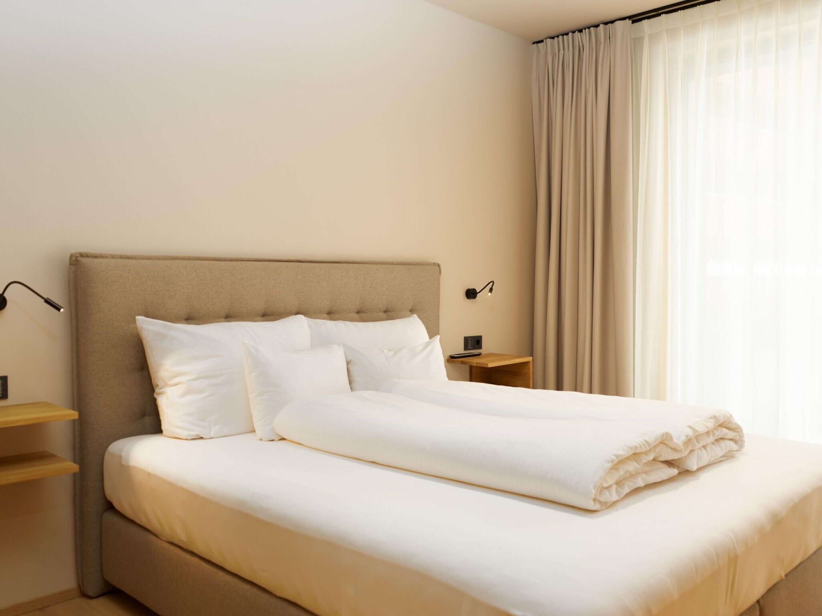 Una cama bien tendida con un cabecero color beige y ropa de cama blanca, una mesita de noche con una lámpara, y una ventana con cortinas de gasa.