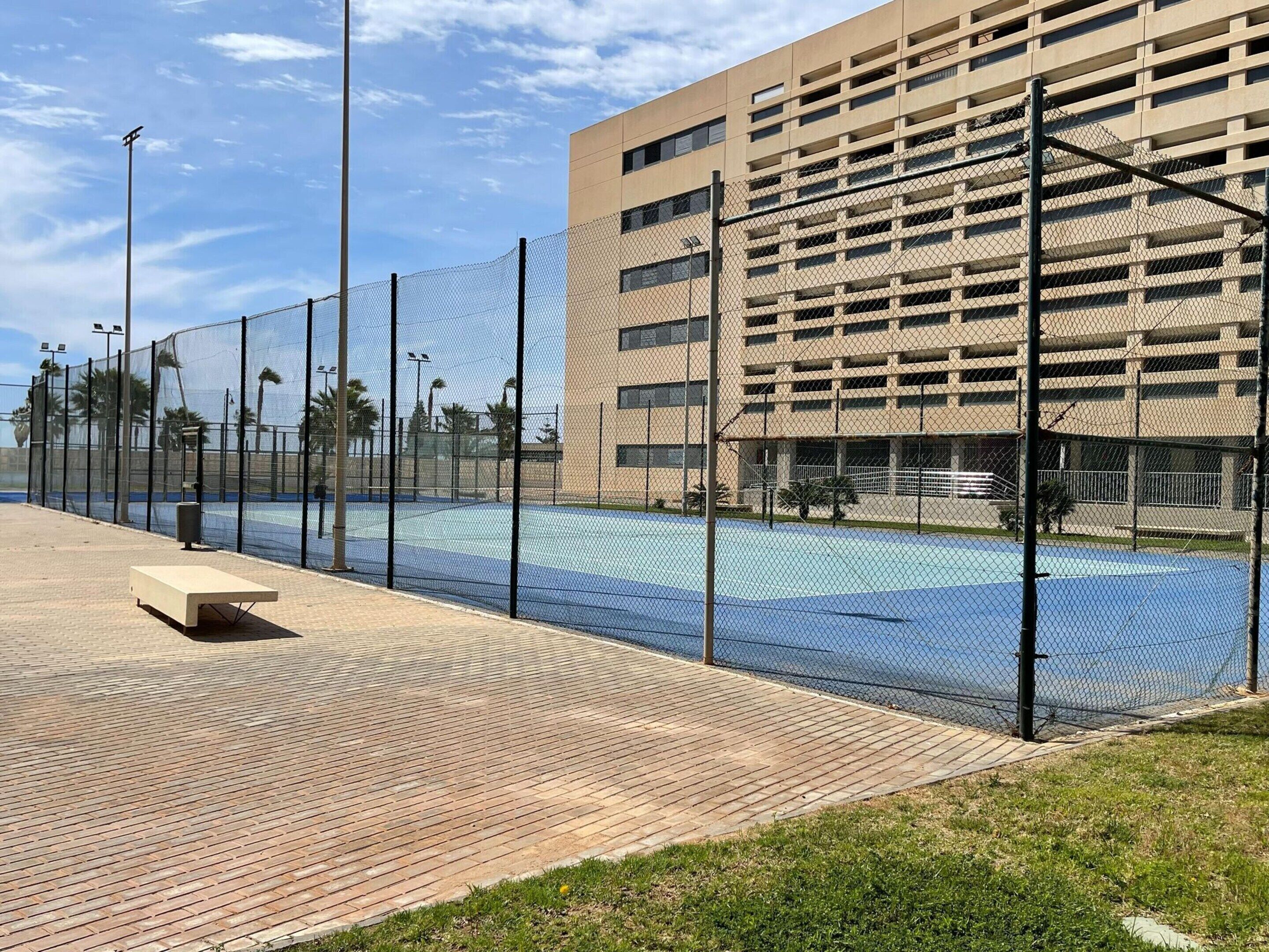 A tennis court with a chain-link fence, a bench, and a building with multiple floors in the background.