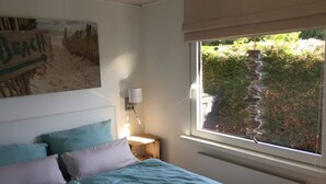 Un dormitorio con cama, almohadas, una mesita de noche, una lámpara, una ventana con vista a follaje y una pared con un letrero de playa.