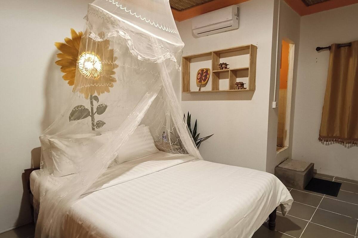 A bed with a white canopy, a wall-mounted air conditioner, a small shelf with decorative items, and a window with curtains.