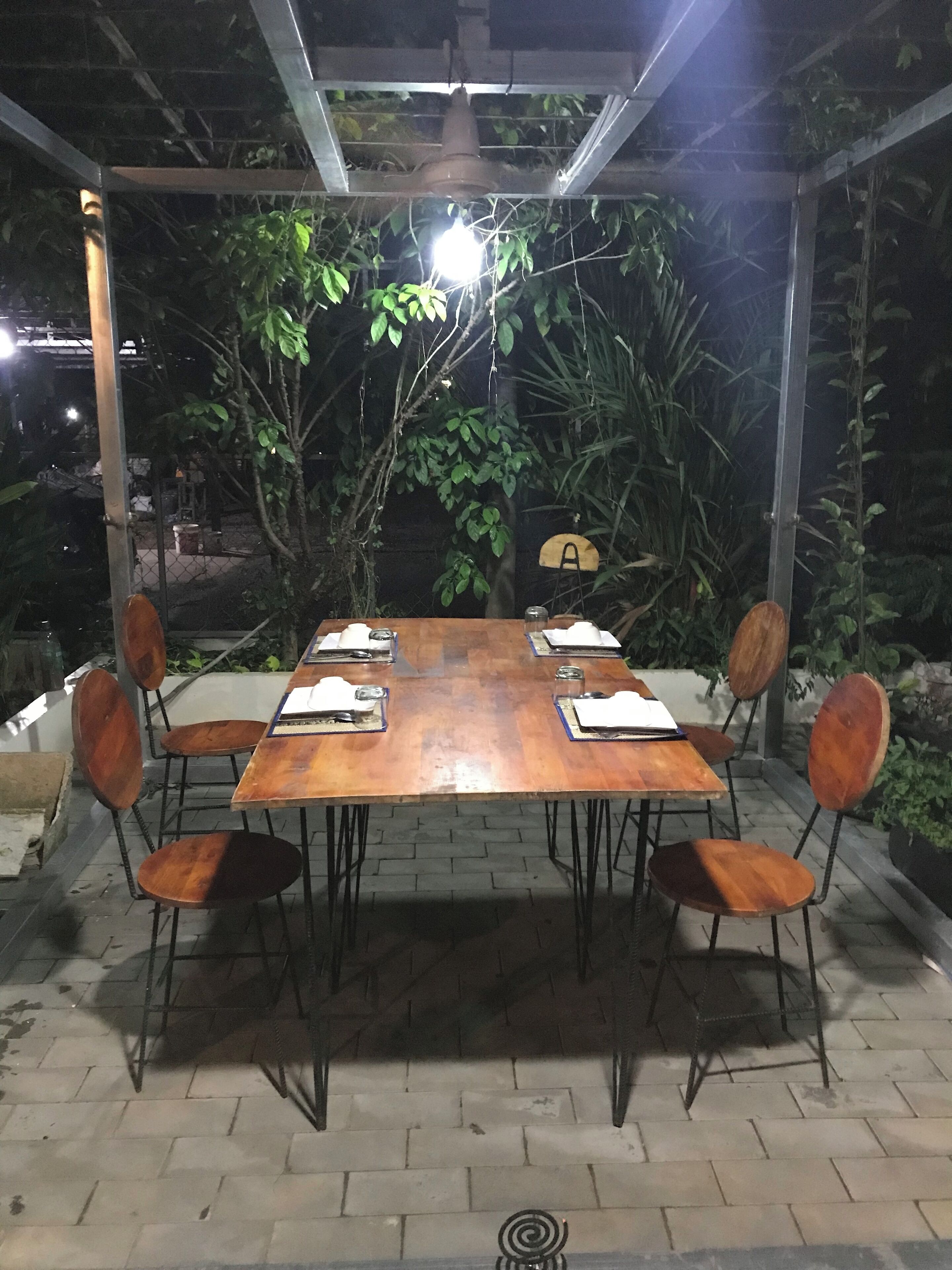 Un área de comedor al aire libre con mesa y sillas de madera, rodeada de follaje y una pérgola de metal.